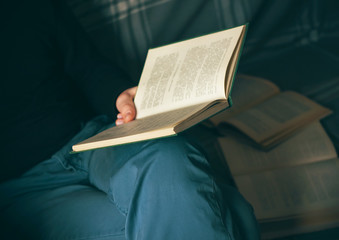 A man in blue pants and a black hoodie sits on a couch with open books scattered around, reading an encyclopedia.