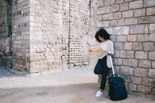 Black tourist with suitcase reading map near old building