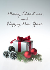 Vertical christmas composition with a gift wrapped with red ribbon, silver cones, red baubles and a fir tree branch - isolated on white background with greeting text