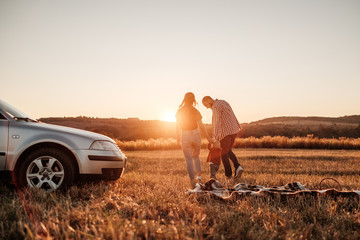 Happy Young Family Mom and Dad with Their Little Son Enjoying Summer Weekend Picnic on the Car Outside the City in the Field at Sunny Day Sunset, Vacation and Road Trip Concept