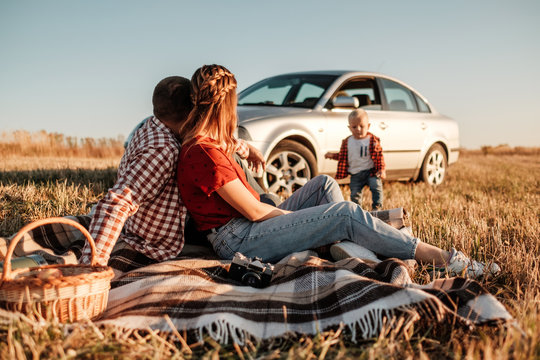 Happy Young Family Mom and Dad with Their Little Son Enjoying Summer Weekend Picnic Sitting on the Plaid Near the Car Outside the City in the Field at Sunny Day Sunset, Vacation and Road Trip Concept