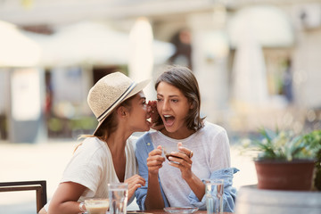  Two beautiful caucasian girls sitting in cafe outside and telling secrets. On table are cups of coffee and grasses with water. Summer time.