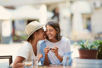  Two beautiful caucasian girls sitting in cafe outside and telling secrets. On table are cups of coffee and grasses with water. Summer time.