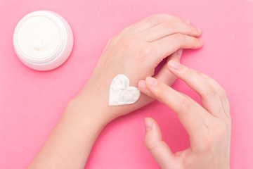 Hands of a beautiful well-groomed woman with a cream jar on a pink textural background. Moisturizer for clean and soft skin in the winter. The heart shape is made of cream.
