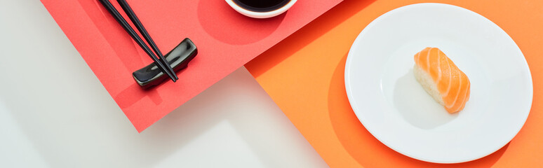 fresh nigiri with salmon near soy sauce and chopsticks on red, orange, white surface, panoramic shot