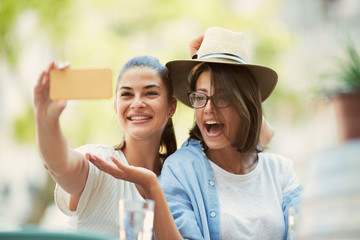 Obraz premium Two beautiful caucasian girlfriends sitting in cafe outdoors and taking selfie.