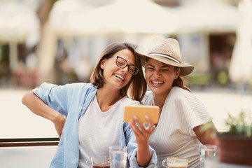 Two beautiful caucasian female friends sitting in cafe, looking at smartphone and laughing.