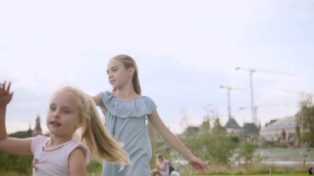 Two adorable long-hair sisters 9 and 5 years old in blue and rose dresses dancing on meadow in Moscow downtown. Kremlin and other people are in defocus on a background. Slow motion, cloudy summer day