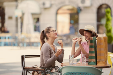 Two cheerful caucasian best female friends enjoying beautiful summer day at coffee shop. On table are shopping bags, water and coffee.