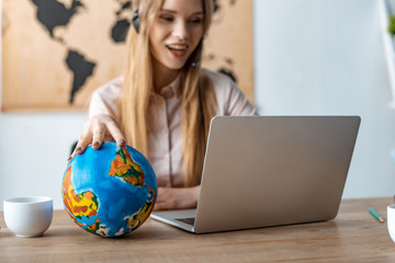 selective focus of smiling travel agent touching globe while looking at laptop