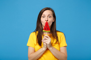 Amazed young brunette woman girl in yellow t-shirt posing isolated on bright blue background, studio portrait. People lifestyle concept. Mock up copy space. Covering mouth with watermelon lollipop.