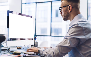 Young businessman typing on computer with blank screen in workspace