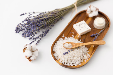 beauty and wellness concept - sea salt with spoon, soap, moisturizer, lavender and cotton flowers on wooden tray