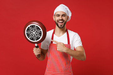 Funny young male chef cook or baker man in striped apron white t-shirt toque chefs hat posing isolated on red background. Cooking food concept. Mock up copy space. Pointing index finger on frying pan.