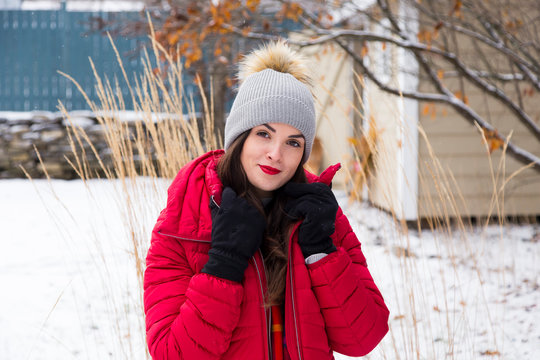 Medium portrait of beautiful smiling young brunette holding the lapels of her puffy red winter coat while standing in garden with ornamental grasses and shed in soft focus background