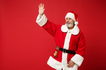 Fototapeta na wymiar Elderly gray-haired mustache bearded Santa man in Christmas hat posing isolated on red background. New Year 2020 celebration concept. Mock up copy space. Waving, greeting with hand as notices someone.