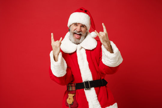 Elderly gray-haired mustache bearded Santa man in Christmas hat posing isolated on red background. Happy New Year 2020 celebration holiday concept. Mock up copy space. Depicting heavy metal rock sign.