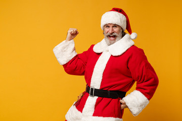 Fototapeta na wymiar Happy elderly gray-haired mustache bearded Santa man in Christmas hat posing isolated on yellow background. Happy New Year 2020 celebration concept. Mock up copy space. Clenching fists like winner.