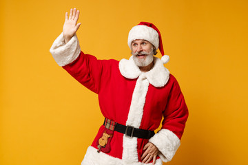 Fototapeta na wymiar Elderly gray-haired mustache bearded Santa man in Christmas hat posing isolated on yellow background. New Year 2020 celebration concept. Mock up copy space. Waving greet with hand as notices someone.