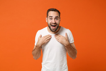 Surprised young man in casual white t-shirt posing isolated on bright orange wall background, studio portrait. People sincere emotions lifestyle concept. Mock up copy space. Holding hands on chest.