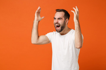 Angry young man in casual white t-shirt posing isolated on bright orange wall background studio portrait. People sincere emotions lifestyle concept. Mock up copy space. Screaming, spreading hands.