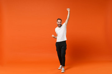 Fototapeta na wymiar Happy young man in casual white t-shirt posing isolated on orange wall background, studio portrait. People sincere emotions lifestyle concept. Mock up copy space. Rising hands, doing winner gesture.