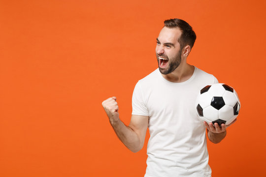 Joyful young man in casual white t-shirt posing isolated on orange background studio portrait. People sincere emotions lifestyle concept. Mock up copy space. Holding soccer ball doing winner gesture.