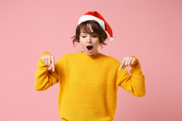 Shocked young brunette woman Santa girl in yellow sweater Christmas hat posing isolated on pink background. New Year 2020 celebration holiday concept. Mock up copy space. Pointing index fingers down.