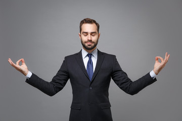 Young business man in classic black suit shirt tie posing isolated on grey background. Achievement career wealth business concept. Mock up copy space. Hold hands in yoga gesture, relaxing meditating.