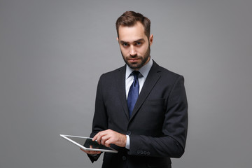 Serious young business man in classic black suit shirt tie posing isolated on grey background. Achievement career wealth business concept. Mock up copy space. Holding and using tablet pc computer.