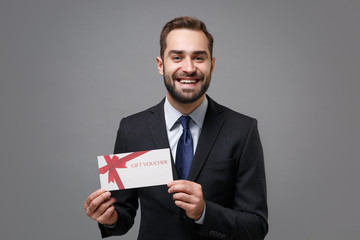 Smiling young business man in classic black suit shirt tie posing isolated on grey wall background in studio. Achievement career wealth business concept. Mock up copy space. Holding gift certificate.