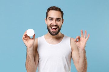 Bearded young man 20s years old applying moisturizer facial cream from container isolated on blue...