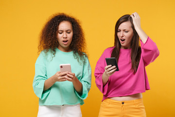 Two displeased women friends european african american girls in pink green clothes posing isolated on yellow background. People lifestyle concept. Mock up copy space. Use mobile phone, typing message.