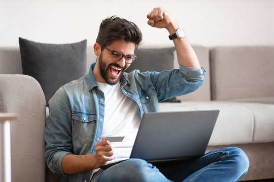 Handsome caucasian hipster holding laptop in lap, using credit card and cheering while sitting on floor in living room.