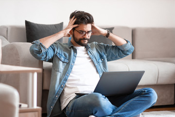 Annoyed caucasian young bearded man with eyeglasses and dressed casual sitting on floor with laptop in lap, holding head and getting nervous about situation on his bank account.