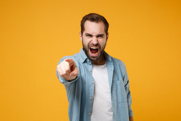 Angry young bearded man in casual blue shirt posing isolated on yellow orange background in studio. People lifestyle concept. Mock up copy space. Screaming swearing, pointing index finger on camera.