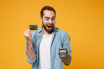 Shocked young man in blue shirt posing isolated on yellow orange background. People lifestyle concept. Mock up copy space. Hold wireless bank payment terminal to process acquire credit card payments.