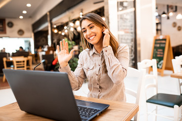 Happy beautiful caucasian brunette young woman waving hello during video call on her laptop. She is cheerful, smiling, enjoying in communication with her friends while sitting in a coffee shop.