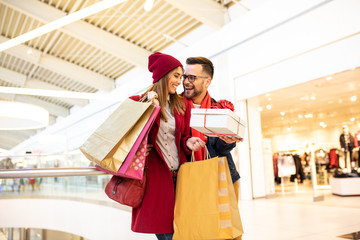 Cheerful young caucasian couple smiling, holding each other and carrying bags after shopping. Young woman is surprised by the gift her boyfriend gave her. They are standing in the shopping mall.