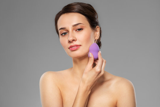 beauty, people and skincare concept - young woman applying foundation with make up blending sponge over grey background