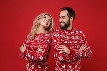 Young couple guy girl in Christmas sweaters posing isolated on red background. Happy New Year 2020 celebration holiday party concept. Mock up copy space. Holding hands crossed, looking at each other.