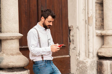 Young handsome caucasian hipster standing near rustic door and typing message on smart phone.