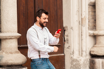 Young handsome smiling caucasian hipster standing near rustic door and reading message on smart phone.