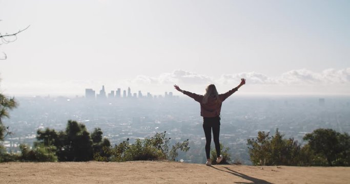 A young woman walking to the edge of a cliff with a view of the city of Los Angeles.