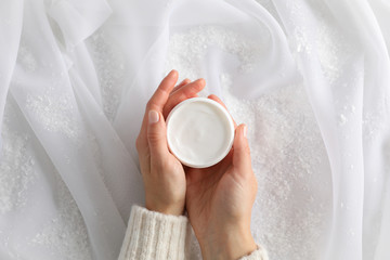 Woman holding jar of winter cream for skin, decorative snow on white fabric background, top view. Space for text