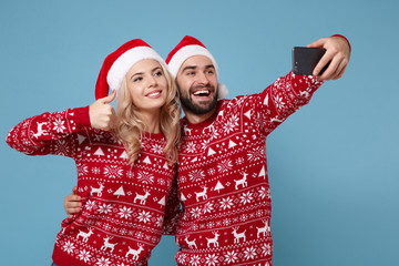 Young couple guy girl in Christmas sweater Santa hat posing isolated on blue wall background. New Year 2020 celebration concept. Mock up copy space. Doing selfie shot on mobile phone showing thumb up.