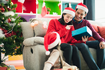 Young Asian family celebration in Christmas day, daughter siting on floor which open give box which smiling felling happy, parent sitting on sofa in living room at home. Merry Xmas happy new year.