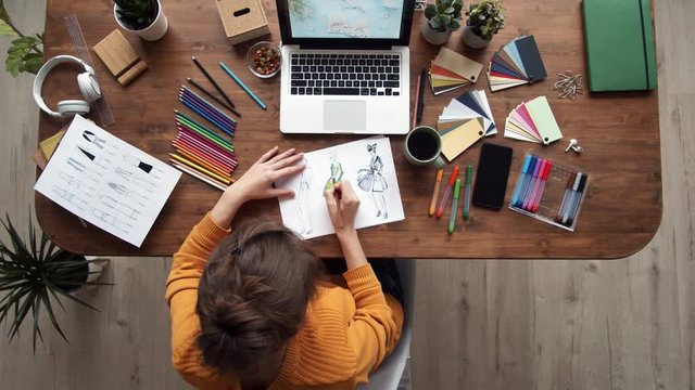 Flat lay of female clothing designer sitting at desk at computer screen and coloring clothes sketches using colour pencils and felt-tip pens