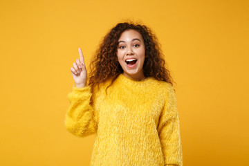 Excited young african american girl in fur sweater posing isolated on yellow orange background, studio portrait. People lifestyle concept. Mock up copy space. Hold index finger up with great new idea.