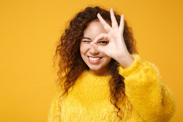 Smiling young african american girl in fur sweater posing isolated on yellow orange wall background. People lifestyle concept. Mock up copy space. Showing OK gesture, imitating glasses or binoculars.